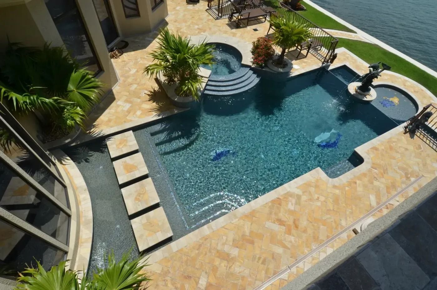 High-angle view of luxury pool with stone and tile surround, near water. Tropical plants, steps, and fountains.