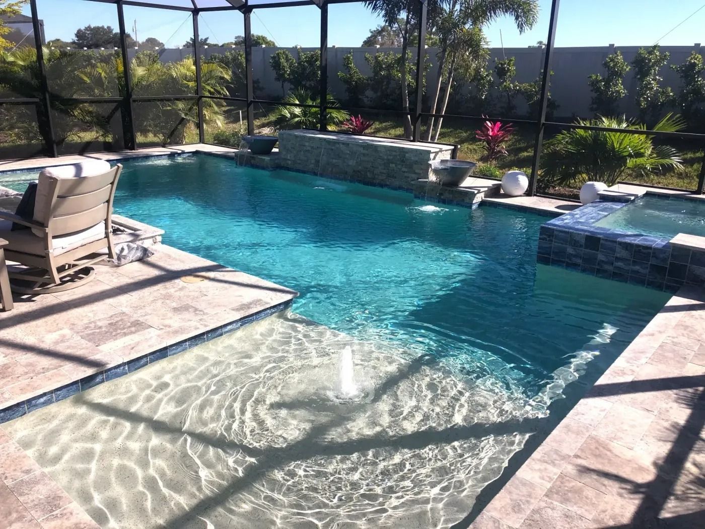 Pool with turquoise water, a waterfall, and attached spa. Tan stone deck, a screened enclosure, and landscaping.
