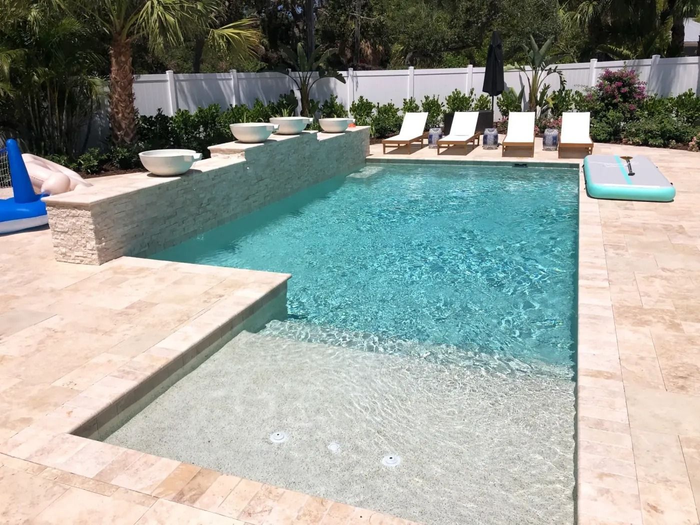 Rectangular pool with travertine tile surround. White wall in the background. Lounge chairs and flotation device nearby.
