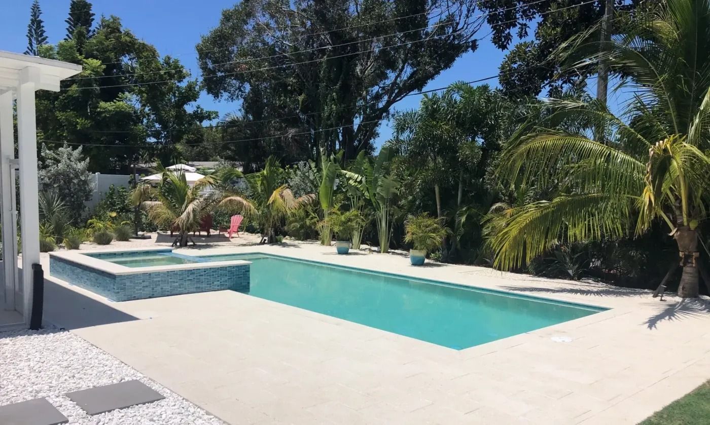 Pool with a hot tub, surrounded by light-colored decking and tropical foliage. Sunny outdoor setting.