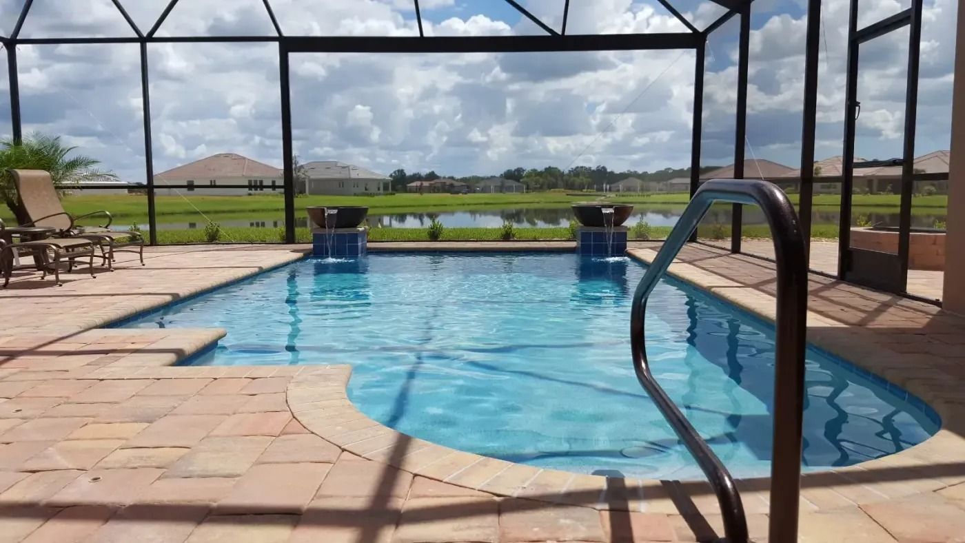 Swimming pool with waterfalls, surrounded by a screened enclosure and brick patio, overlooking a lake and grassy field.