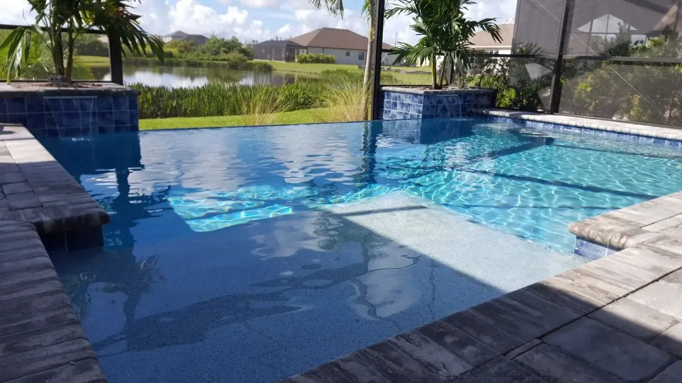 Swimming pool with dark blue tiles, steps, and water reflecting sky, near a lake and houses.
