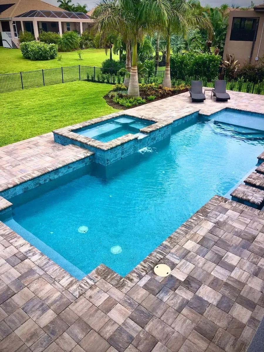 Swimming pool with spa, blue water, stone patio, green lawn, palm trees, two lounge chairs.