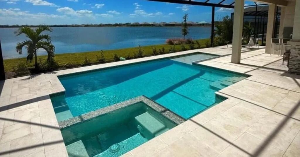 Swimming pool overlooking a lake, teal water, beige patio, palm tree, and pergola.
