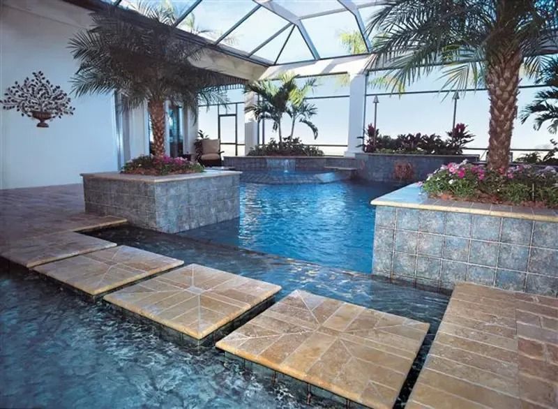 Indoor pool with palms, stone steps, and glass ceiling, surrounded by lush plants and windows.