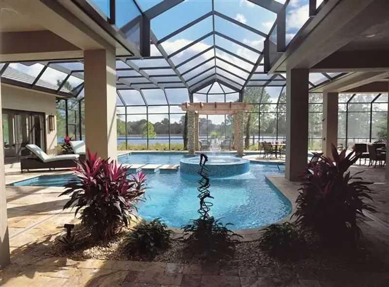 Indoor swimming pool with glass roof, stone patio, plants, and a view of a lake.