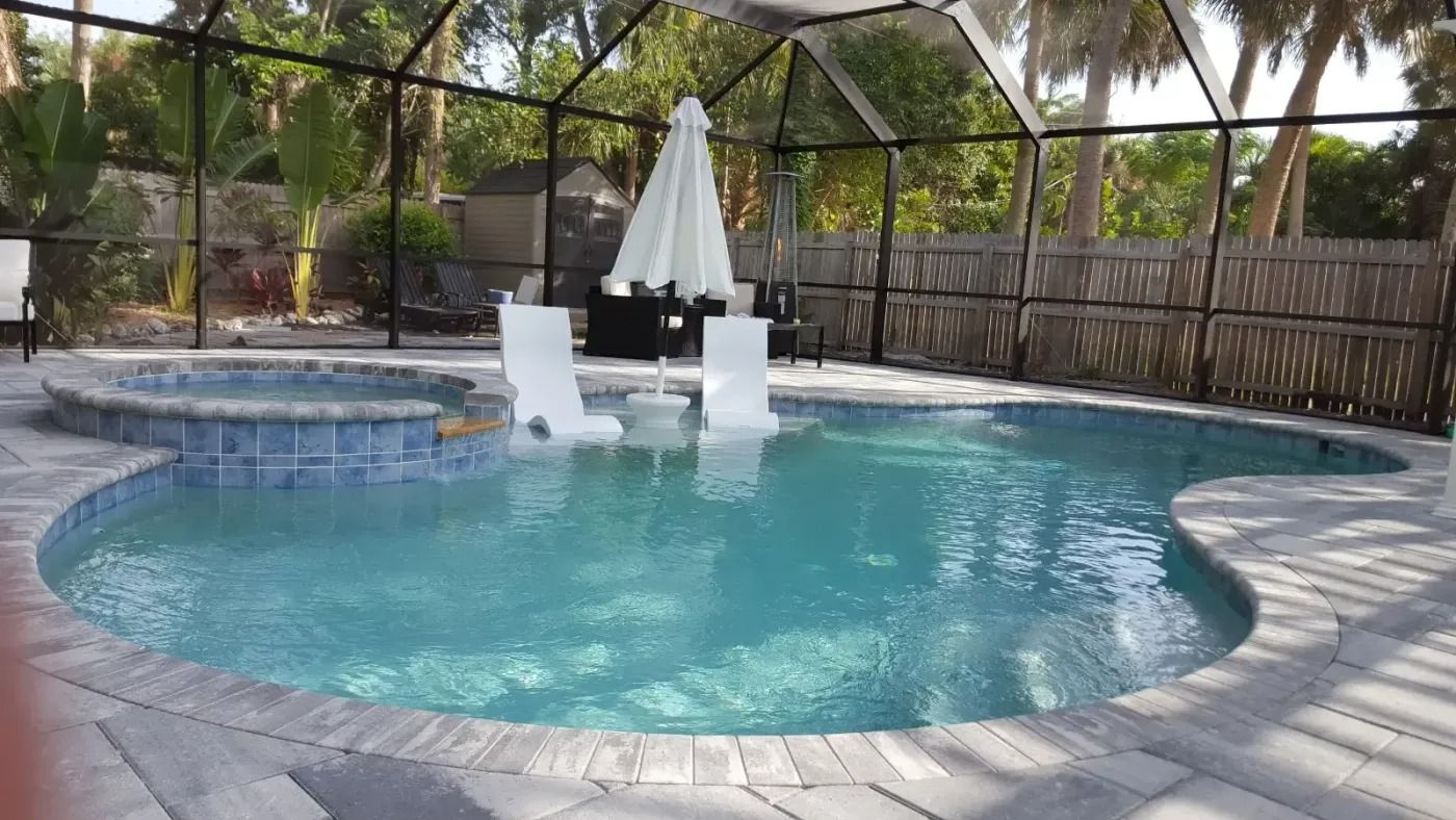 Pool with spa, white chairs, and an umbrella, surrounded by a screen enclosure and wooden fence.
