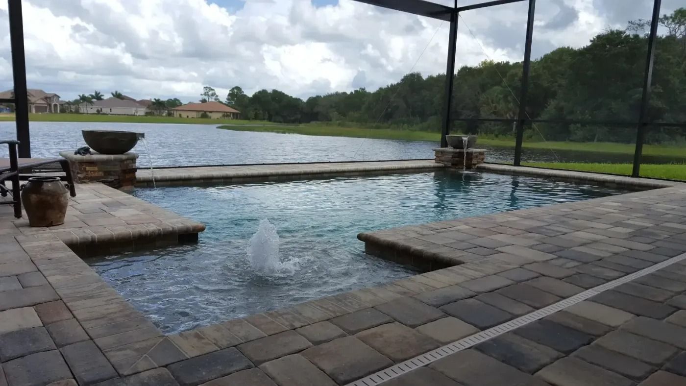 Pool with fountain, surrounded by pavers, overlooking a lake, under a screened enclosure.