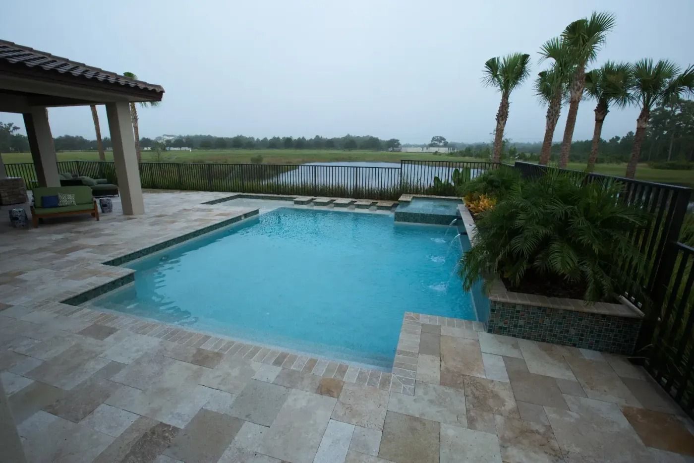 Outdoor pool with light blue water, surrounded by tan stone patio and dark fence, on a cloudy day.