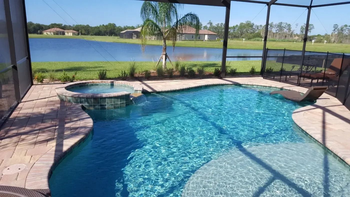 Swimming pool with spa overlooking a lake; patio screen, palm tree, blue water, and green grass.