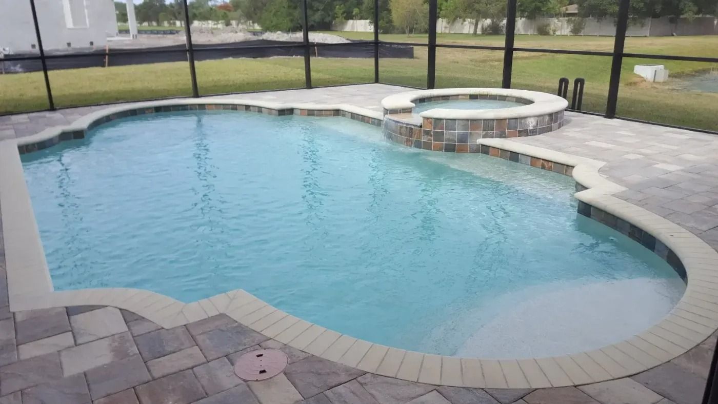 Swimming pool with attached hot tub, surrounded by stone pavers, screened enclosure, and grassy lawn.