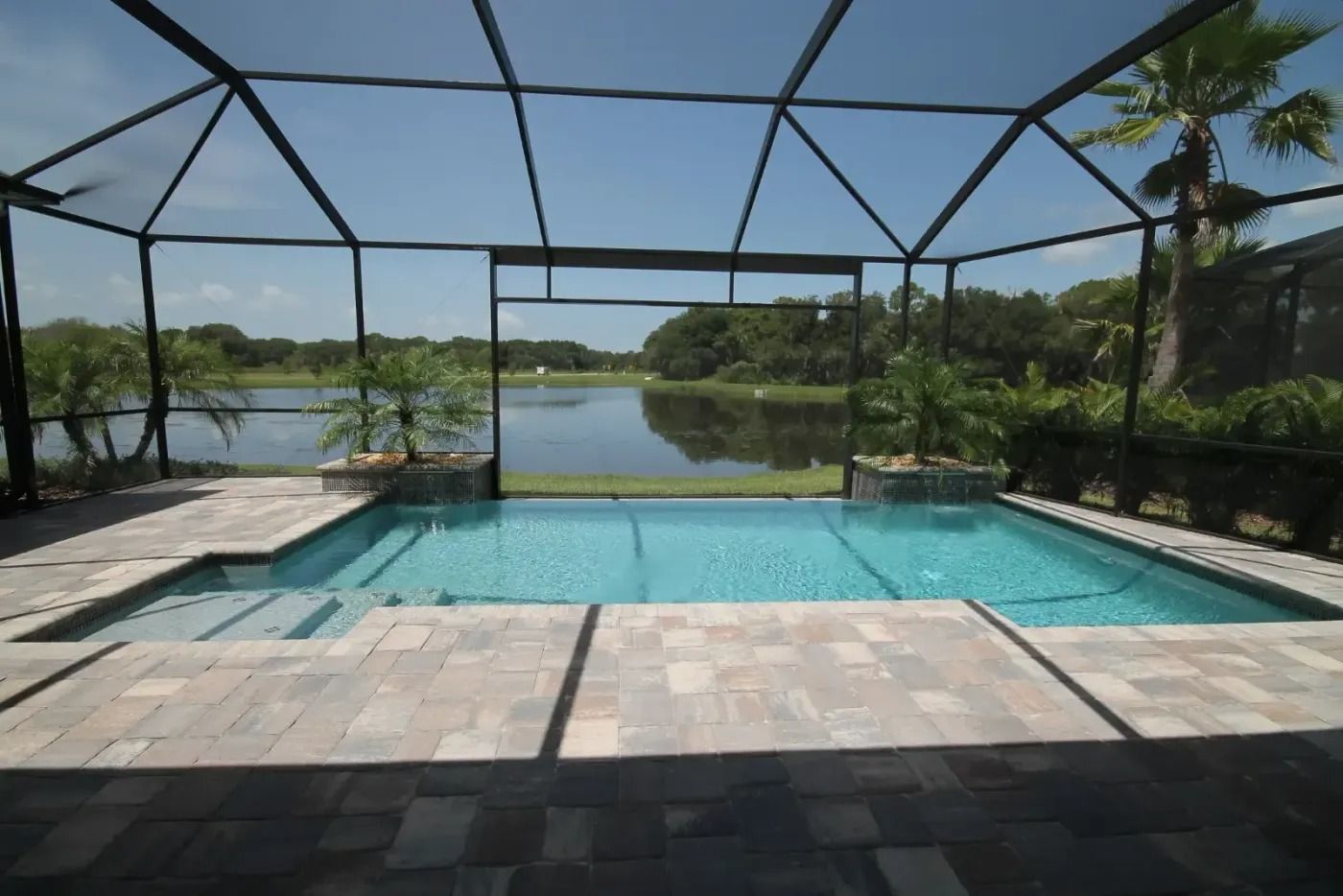 Pool enclosure with pool overlooking a lake, blue water, gray patio, clear sky.
