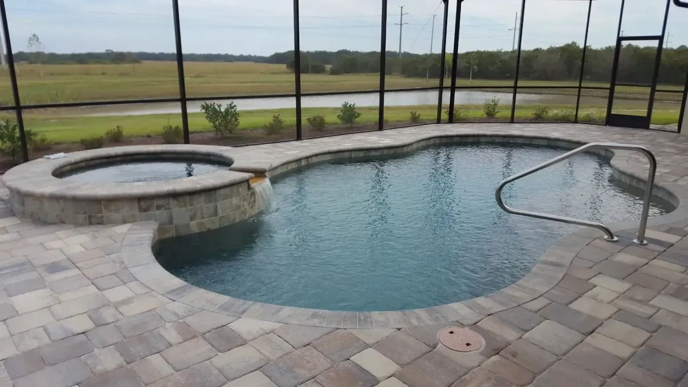 Swimming pool with attached spa, surrounded by pavers and a screen enclosure, set outdoors with a landscape view.