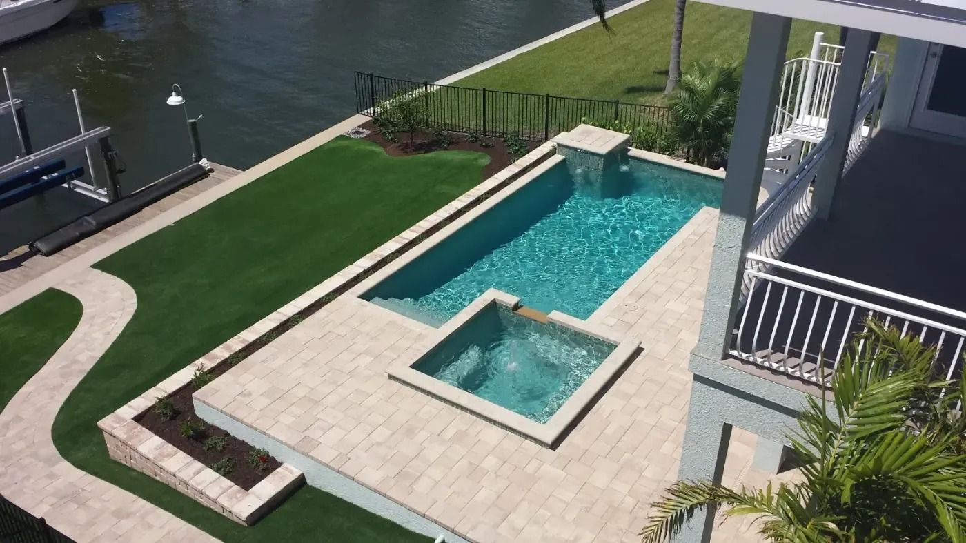 Aerial view of a rectangular pool with a spa, surrounded by green turf and a house overlooking a waterway.