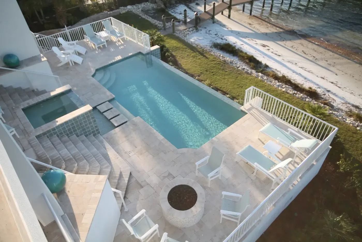 Aerial view of a coastal patio with a pool, hot tub, and fire pit; white furniture and railing.