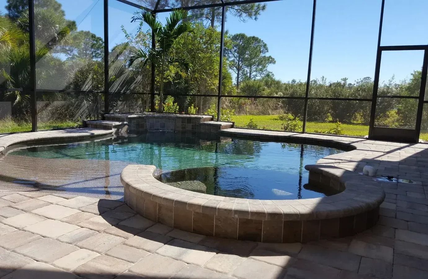 Pool area with dark screen enclosure, stone patio and pool, and natural background.