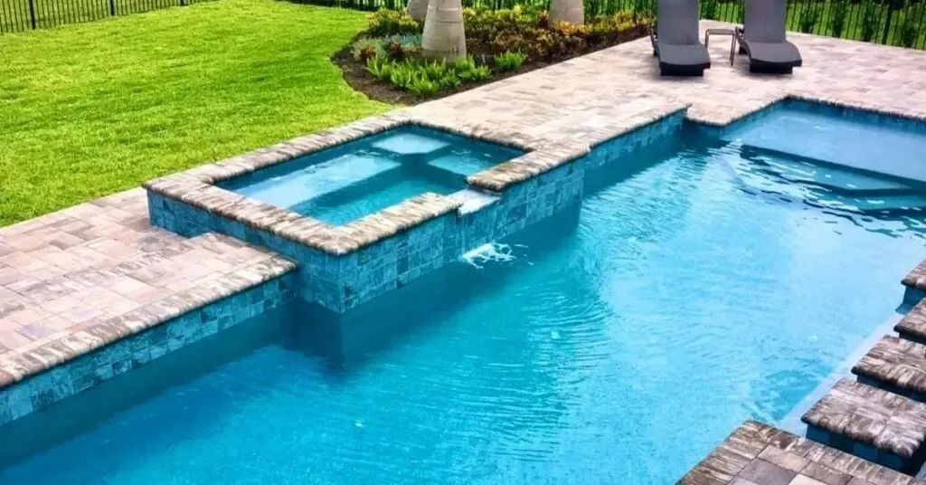 Blue tiled pool with attached hot tub, set in a paved patio, near green grass.