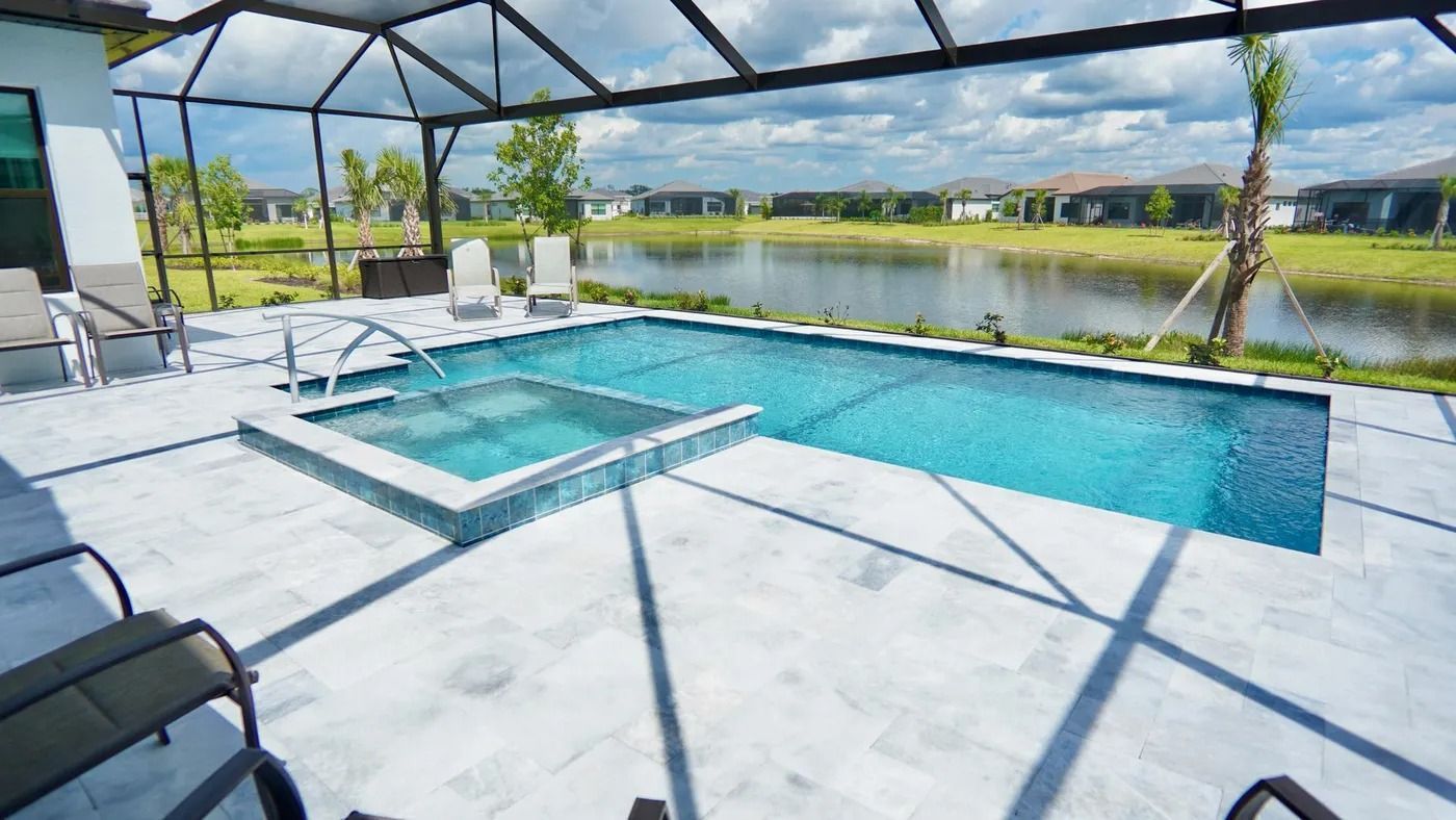 Pool and spa with water surrounded by light colored tiles, next to a lake under a screened enclosure.
