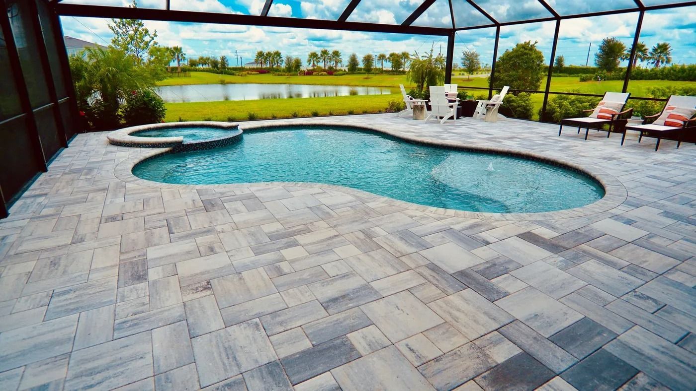 Poolside patio with pool and hot tub overlooking a lake. Gray pavers, chairs, and screened enclosure.