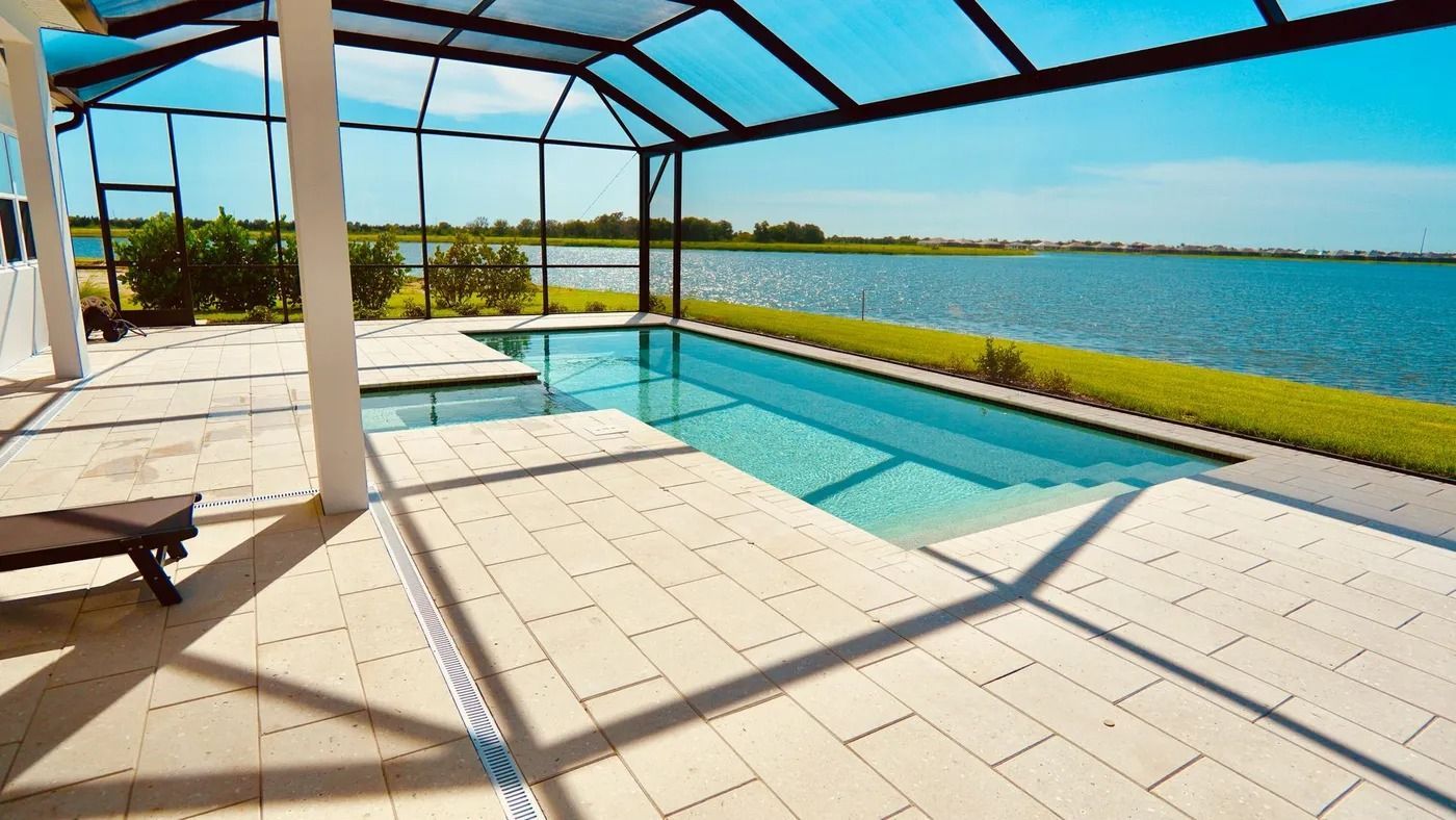Pool with screen enclosure overlooking a lake. Light-colored patio, blue water, and sky.