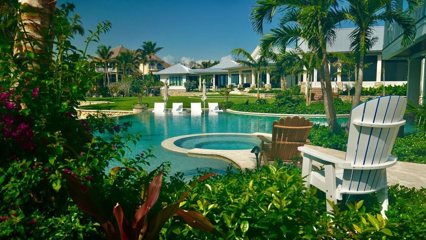 Pool with lounge chairs, lush greenery, and houses under a clear blue sky.