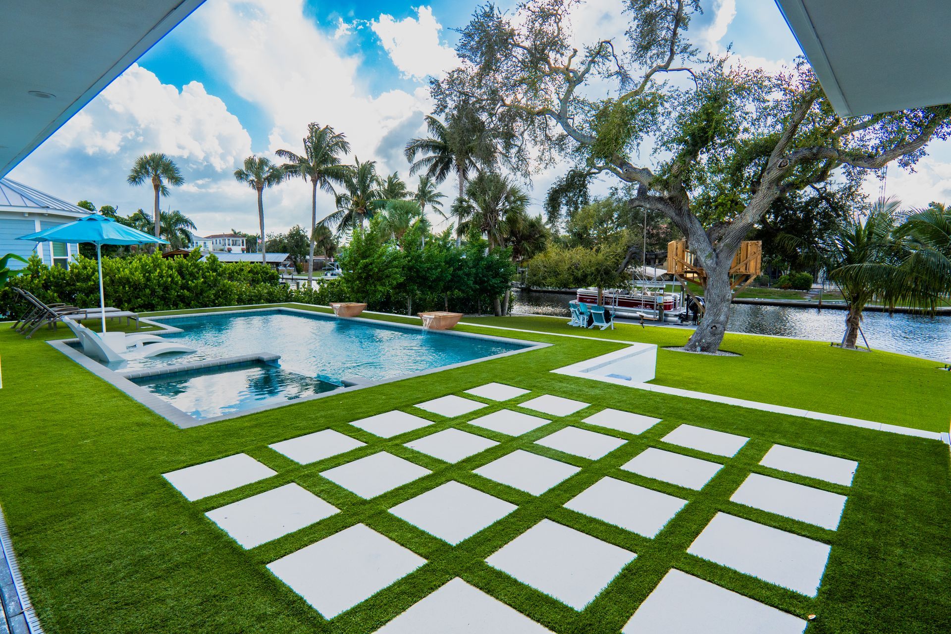 A swimming pool with white paver tiles set into a grassy backyard overlooking a canal with palm trees under a blue sky.