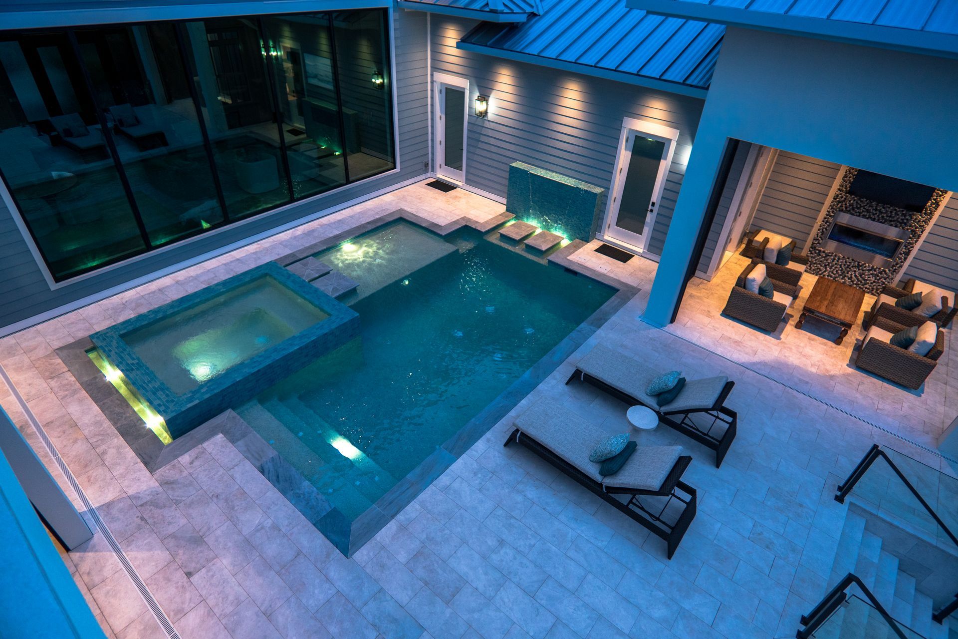 An evening view of a modern luxury backyard featuring a pool, hot tub, lounge chairs, and a covered patio seating area.