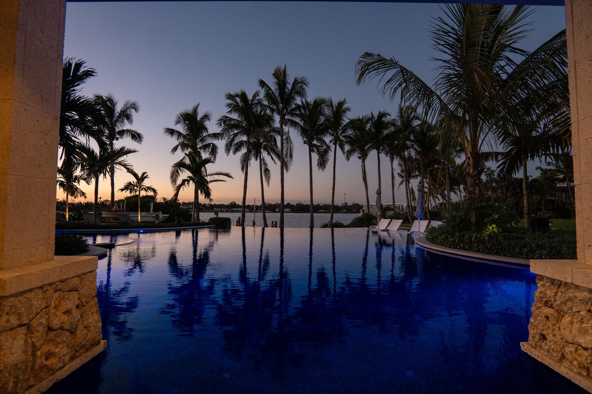 A luxury infinity pool at sunset, reflecting a line of tall palm trees against a soft, twilight sky.