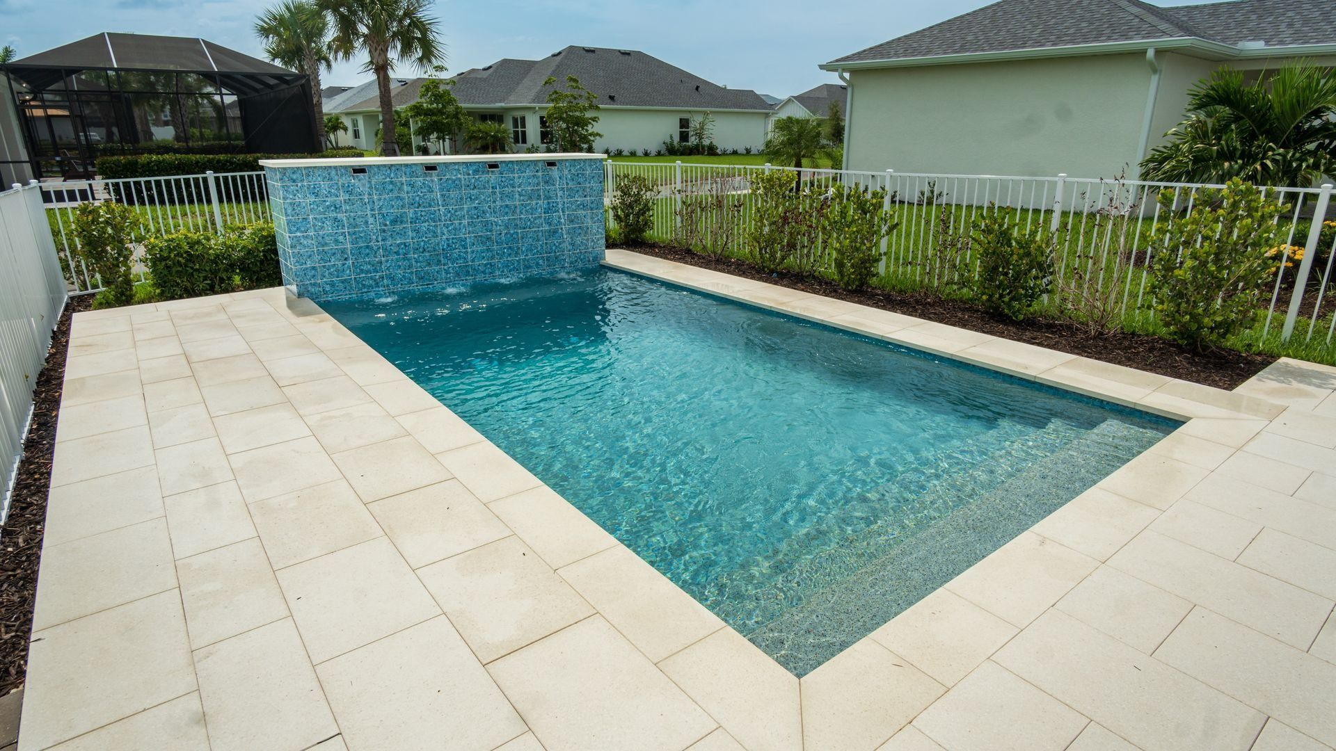 Modern backyard swimming pool with blue water and a tiled patio, surrounded by hedges and suburban homes