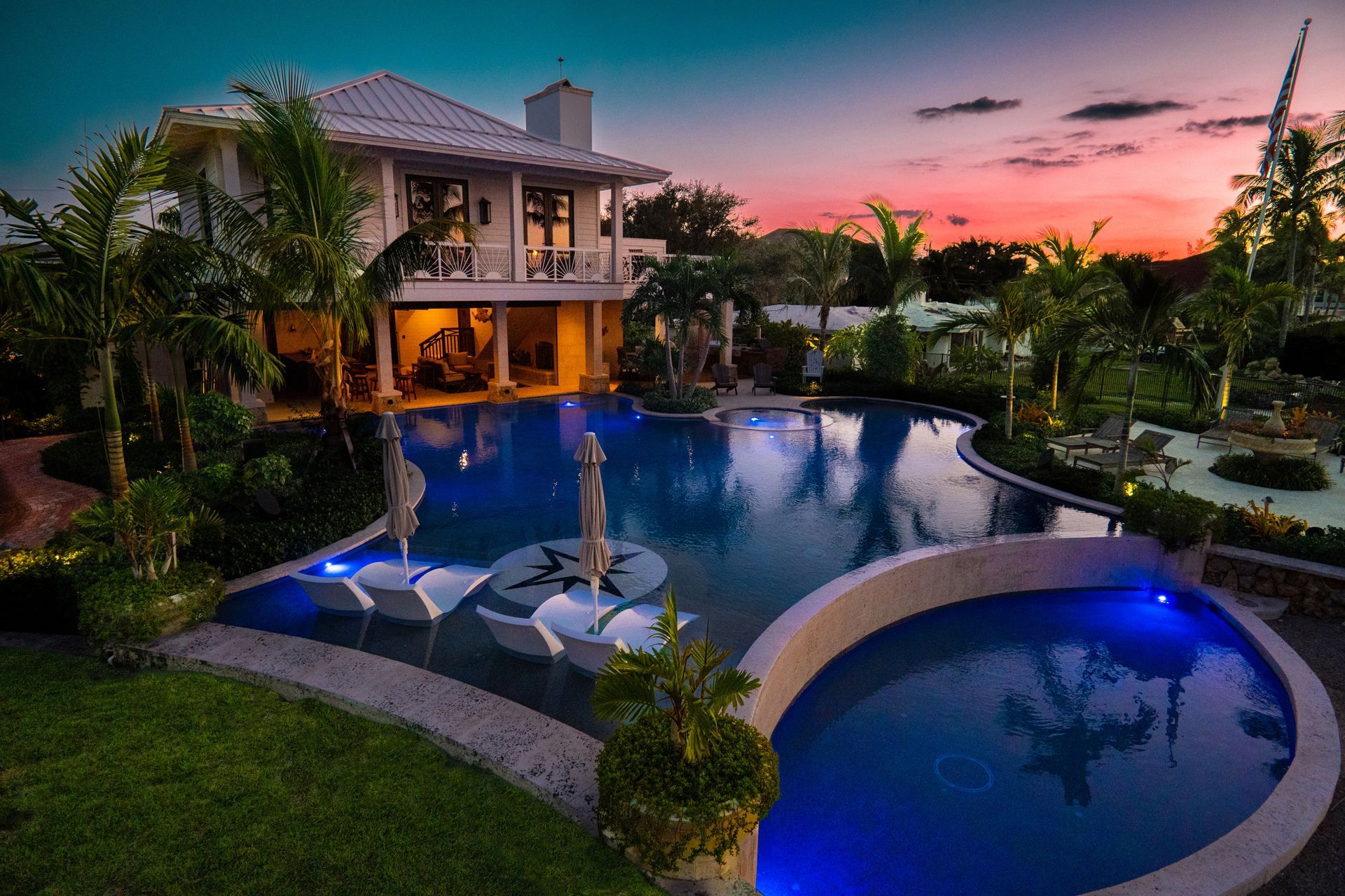 A multi-level home overlooks a glowing pool and hot tub at sunset, surrounded by lush palm trees and landscaped grounds.