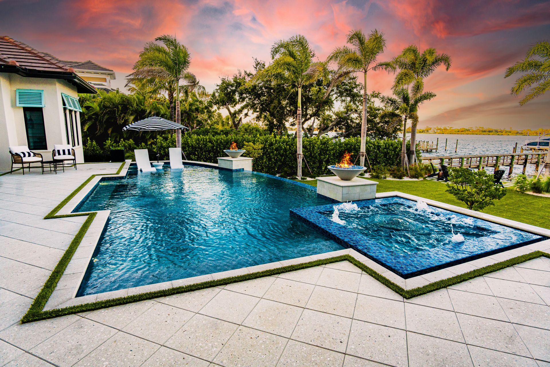An infinity pool and spa overlooking a bay at sunset, featuring stone decking, lounge chairs, and two fire bowls.