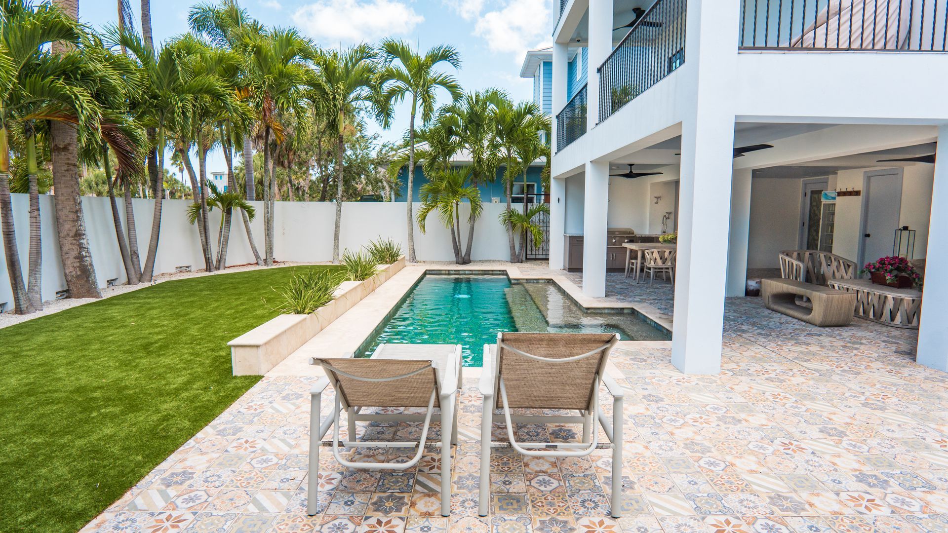 Modern patio with two lounge chairs beside a narrow pool, tropical landscaping, and a white house exterior