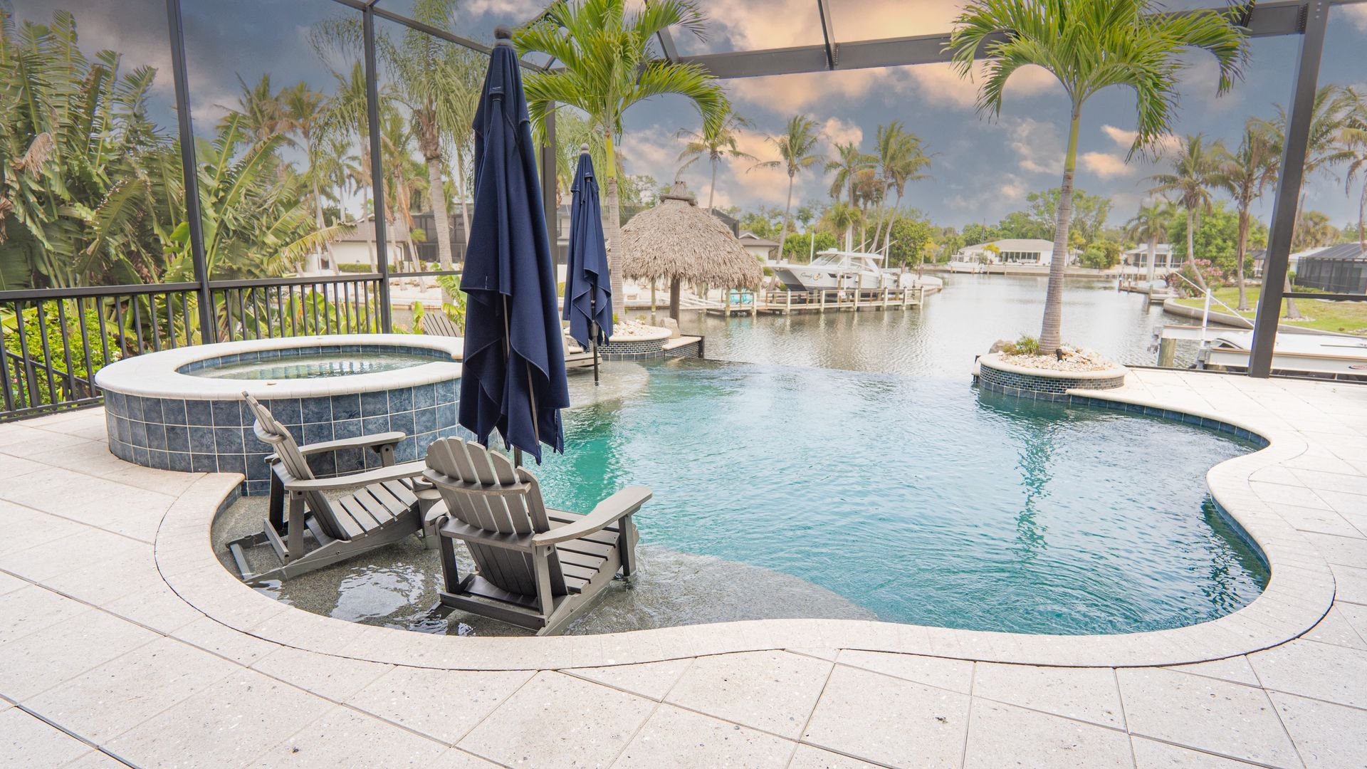 Resort pool with spa, lounge chairs, and umbrellas under a pergola, overlooking water and palm trees
