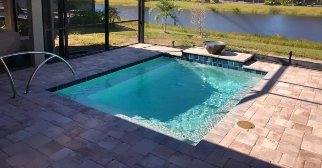 Small, square, turquoise pool with brick patio, near a lake. Handrail and a small waterfall.