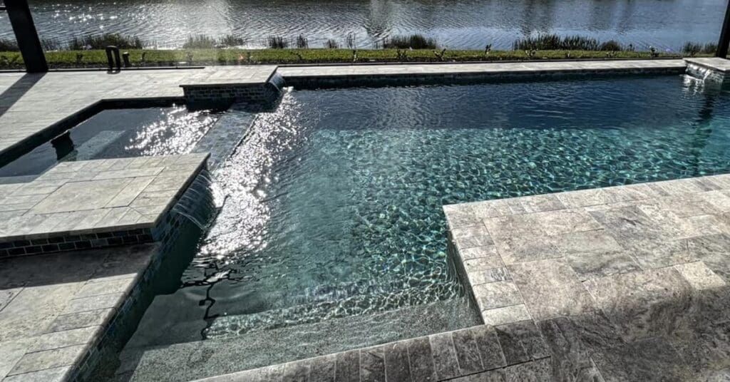 Stone steps lead into a clear, turquoise pool with a waterfall feature overlooking a lake.