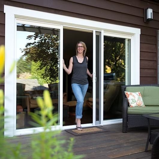 Woman in doorway, holding open sliding glass door. Outside on a deck, with greenery and patio furniture.