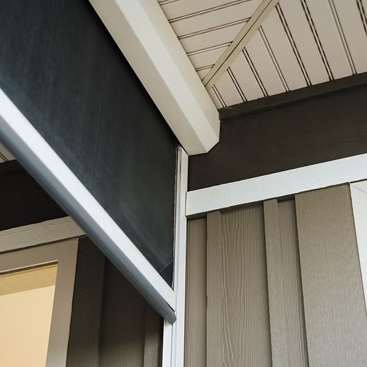 Close-up of a dark grey retractable screen on a porch. The screen is housed in a white frame, with tan siding and white trim visible.