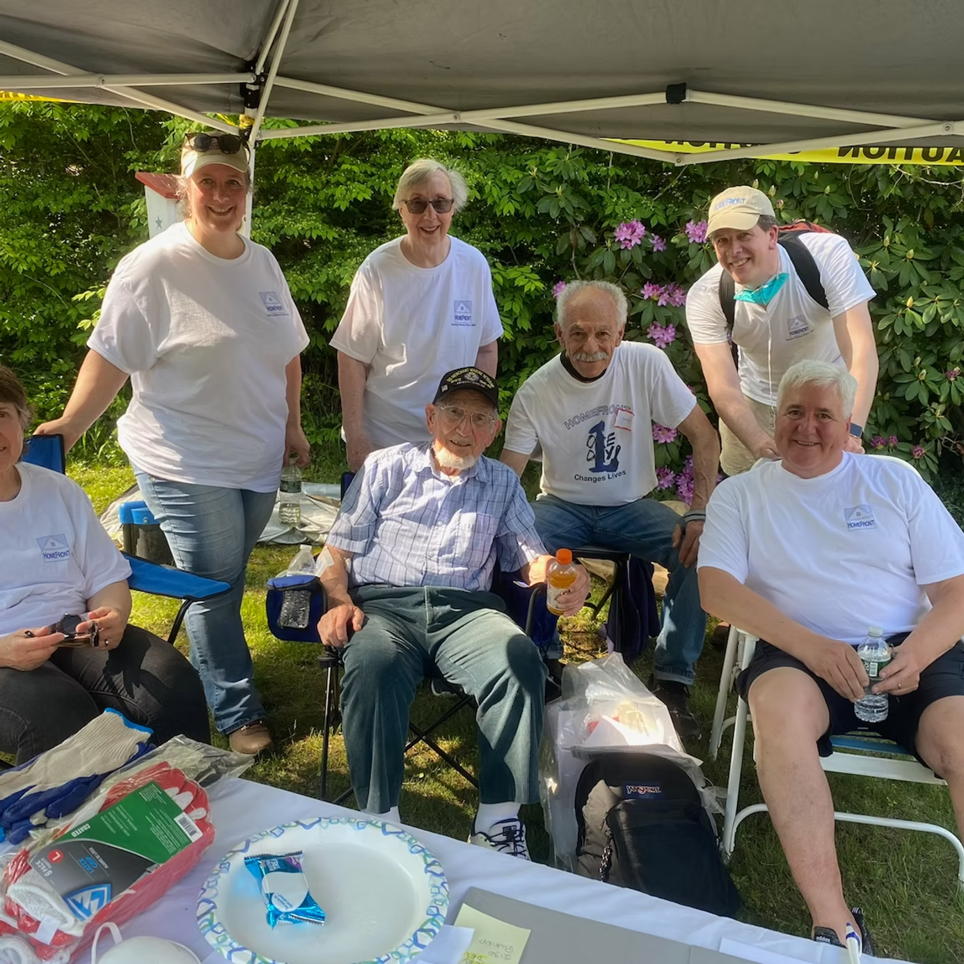 Group of people at an outdoor event, some smiling, gathered around a table with snacks and a seated person.