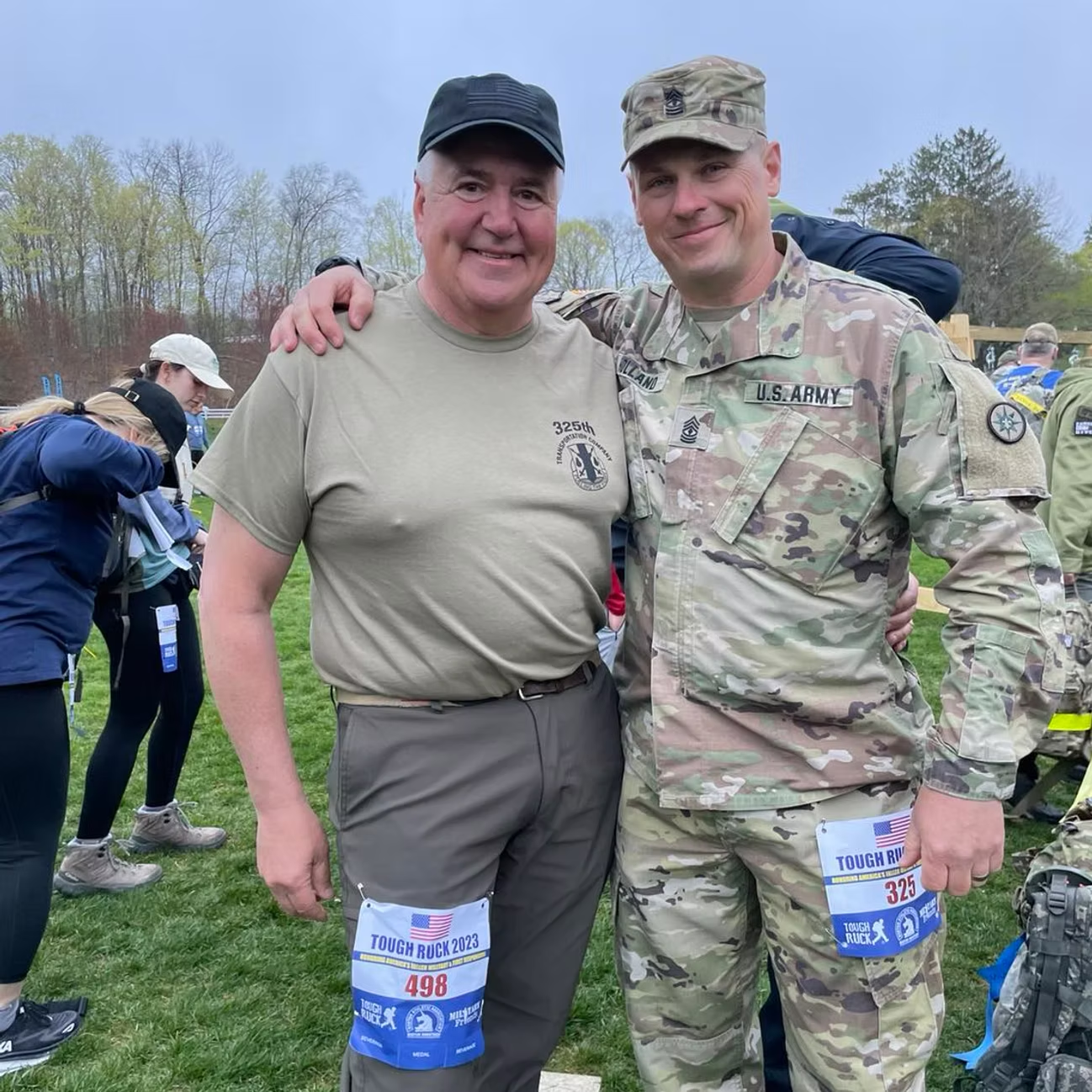 Two men in athletic attire pose for photo outdoors. One in uniform has arm around the other. Both have race bibs attached.