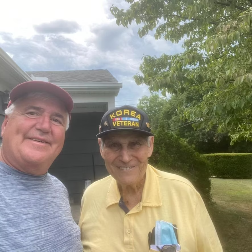 Two men pose outside. One wears a Korean Veteran hat and yellow shirt. The other wears a red cap and gray shirt.