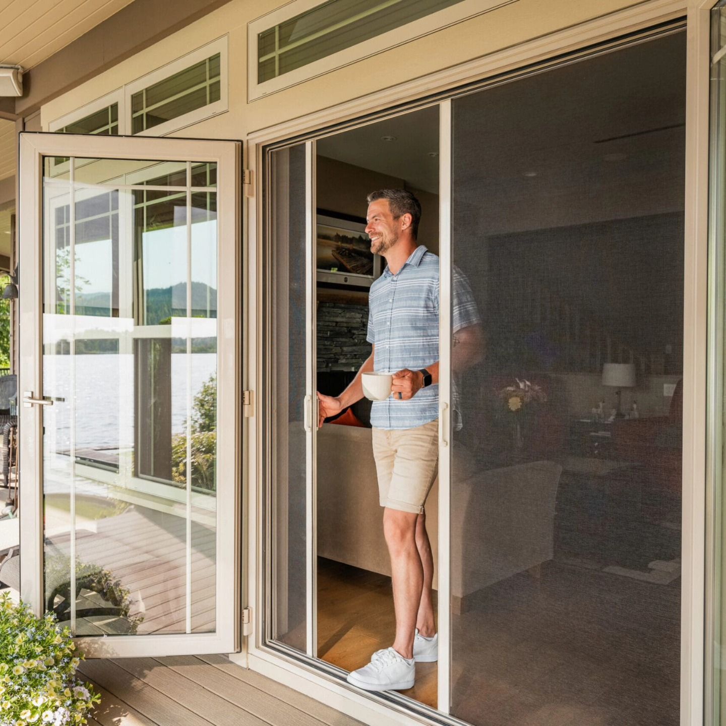 Man holding coffee in screen door doorway, looking outward onto a deck, with lake visible in the background.