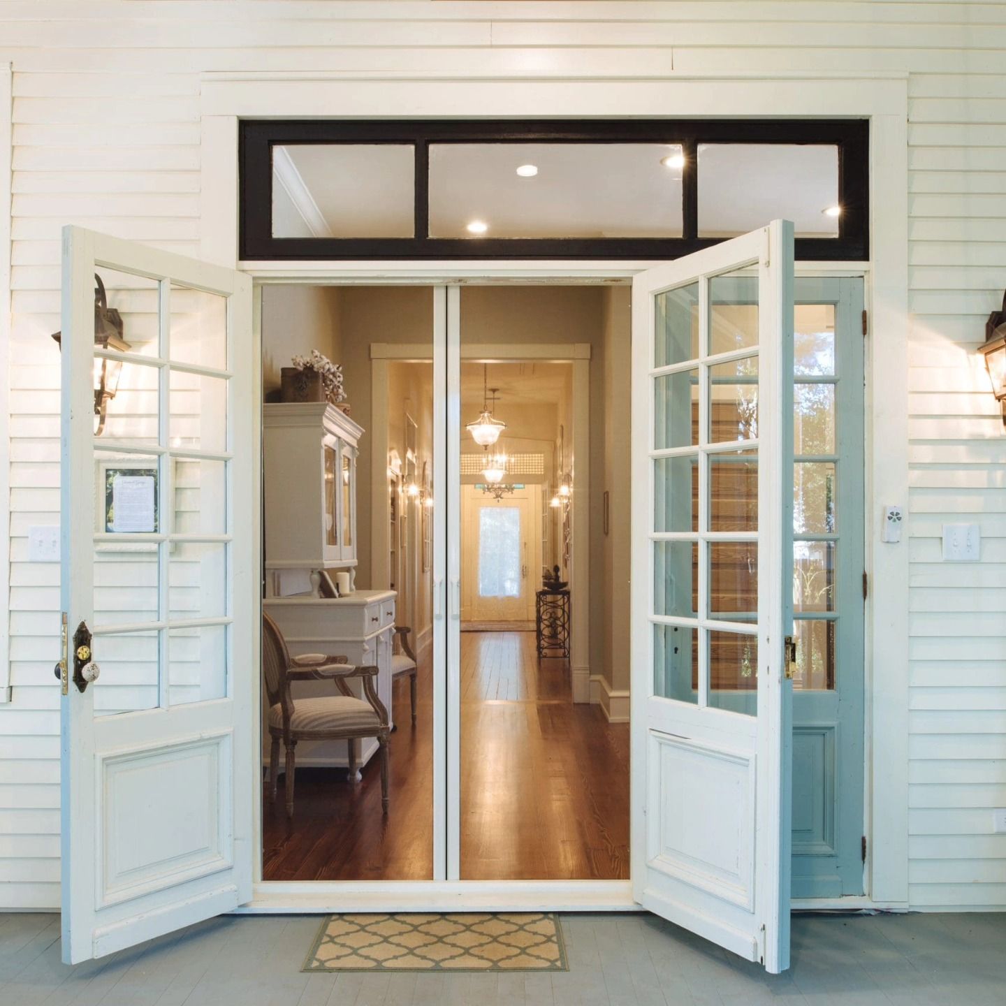 Open white French doors leading into a hallway, flanked by wall sconces, under a black-framed transom.