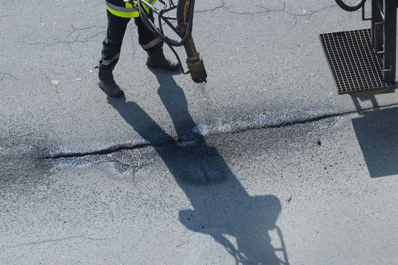 Person in work boots using a tool on cracked pavement, with a long shadow cast on the ground.