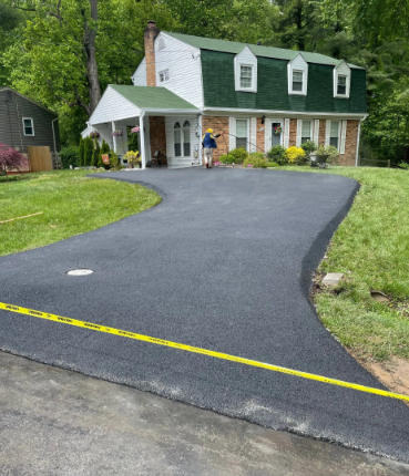 Newly paved asphalt driveway curves toward a two-story brick house with green roof and manicured lawn.