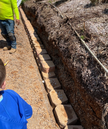 A person builds a retaining wall with blocks in a trench next to a path.