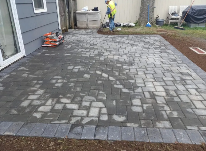 Newly constructed gray brick patio with border; worker in background.