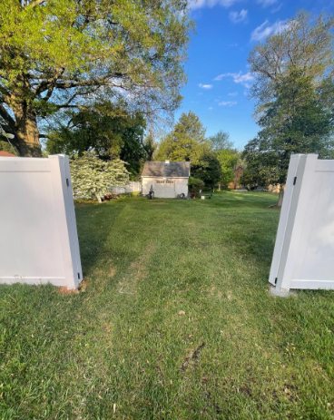 White fence gates open to a grassy yard with a small shed, trees, and blue sky.