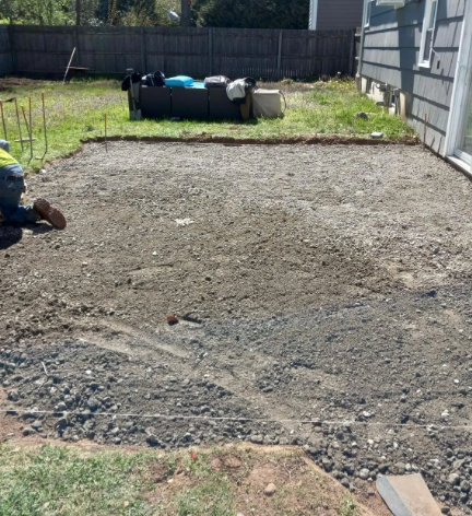Person kneeling, leveling gravel base for a patio. Yard with gray house and wooden fence visible.