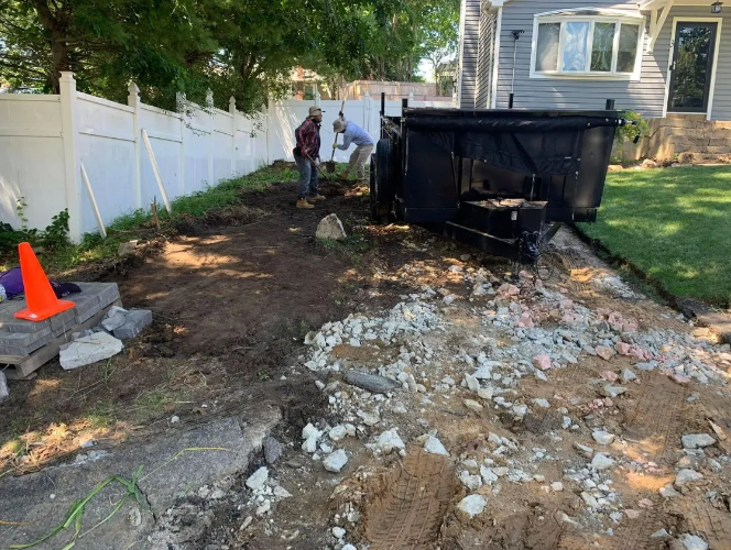 Construction site. Two people work on removing a paved area next to a house with a black dumpster.