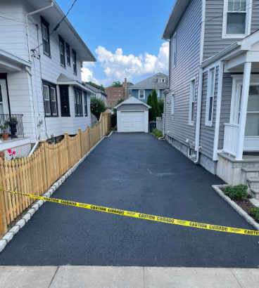 Newly paved asphalt driveway between two houses, with a yellow caution tape in front.